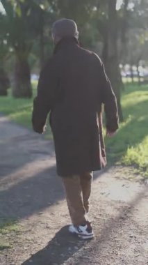 Kahverengi ceketli bir adam güneşli bir günde parktaki palmiye ağaçları arasındaki yolda yürüyor.
