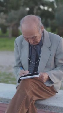 Not defterine yazan yaşlı bir adam parkta bankta oturuyor.