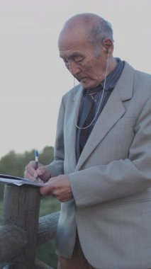 Takım elbiseli ve kulaklıklı kıdemli gazeteci parkta not defterine notlar yazıyor.