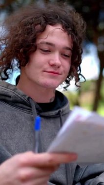 Kıvırcık saçlı öğrenci notları değerlendirir ve not defterine yazar. Parktaki bankta oturur.