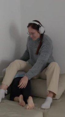 Kulaklık takan mutlu anne, parlak oturma odasındaki rahat bir kanepede neşeli oğlunu gıdıklıyor.