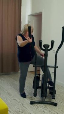 Oturma odasında eliptik eğitmen kullanan kıdemli bir kadın aktif yaşam tarzını sürdürüyor.