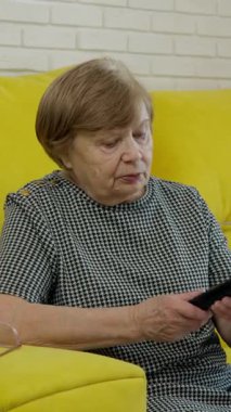 Televizyon izleyen yaşlı bir kadın, sarı kanepede kanal değiştiriyor.