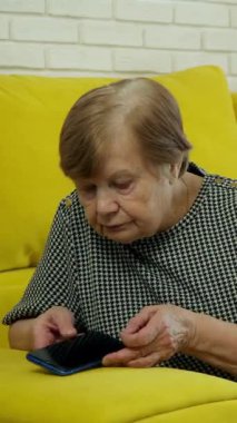 Sarı bir kanepede oturan yaşlı kadın akıllı telefonunu kullanarak bir telefon görüşmesi yapıyor.