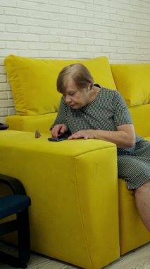 Sarı bir kanepede oturan yaşlı kadın akıllı telefonunu kullanarak bir telefon görüşmesi yapıyor.