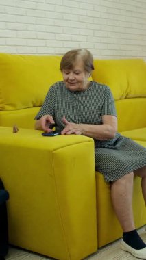 Video görüşmesi için telefonu kullanan mutlu bir emekli kadın, evdeki rahat koltukta oturuyor.