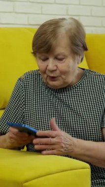 Yaşlı bir kadın sarı bir kanepeye oturur ve ekranda el sallayarak akıllı telefon kullanır.
