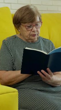 Kıdemli kadın, canlı sarı bir kanepede rahatça otururken kitaptan zevk alıyor.