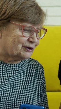 İki yaşlı kadın sarı bir kanepede oturuyor, akıllı telefona bakıyor ve içeriğini tartışıyorlar.