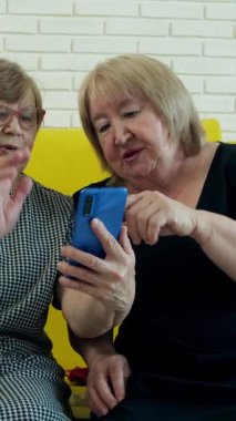 Sarı bir kanepede oturan iki yaşlı kadın video görüşmesi için akıllı telefon kullanıyor.