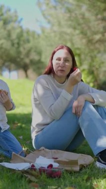 Kadın ve genç çocuk yeşil çimenler ve ağaçlarla çevrili güneşli bir parkta rahatlatıcı bir piknik sırasında taze meyve paylaşıyorlar.