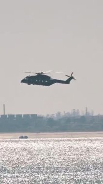 Askeri helikopter arka planda sanayi binaları olan bir kıyı bölgesinin üzerinde uçuyor.