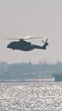 Denizin üzerinde uçan askeri helikopter, bir uçak gemisi ve bir motorlu tekne yakınında.