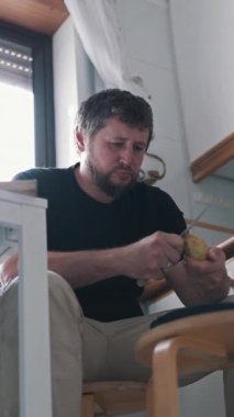 Elmayı dikkatlice soyan bir adam, ev mutfağında bir taburede oturuyor.