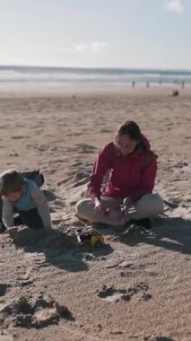Anne ve oğlu, güneşli bir sahilde oyuncaklarla oynayarak kaliteli vakit geçiriyorlar.