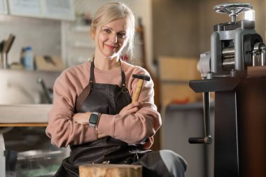 Önlüklü, gülümseyen kadın kuyumcu atölyesinde çekiçle dikiliyor.