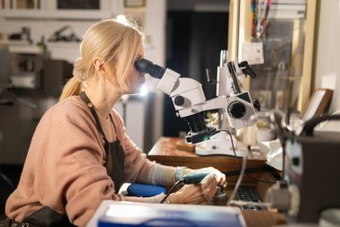 Kadın zanaatkar, atölyesindeki bir mücevhere mikroskop ve döner alet kullanarak konsantre oluyor.