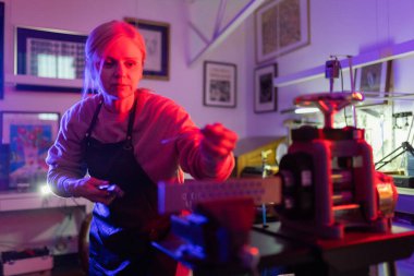 Kadın sanatkar konsantre oluyor, renkli ışıkları olan bir stüdyoda mücevher parçası üzerinde çalışıyor.