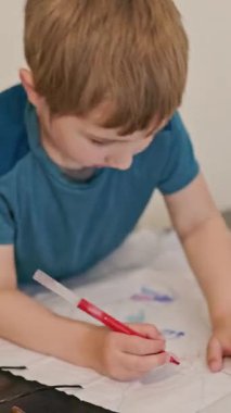 Anaokulu öğrencisi karnının üstüne yatmış, kırmızı kalemle kağıda çizim yapıyor.