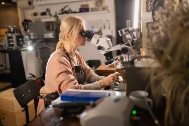 Kusursuz işçiliğe odaklanan bir kadın, stüdyosundaki detayları incelemek için mikroskop kullanıyor.