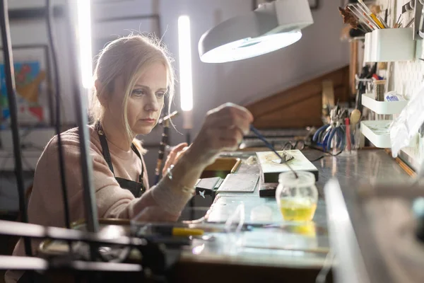 Mücevher, iyi ışıklandırılmış bir stüdyodaki tezgahındaki metal bir nesneye dikkatlice malzeme uyguluyor.