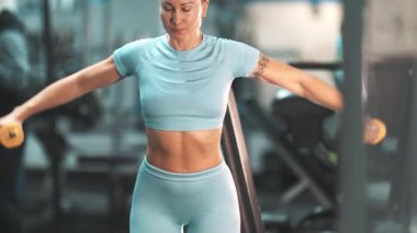 Kas yığını kadın, spor salonunda yoğun bir egzersiz sırasında dambıllarla omuz egzersizi yapıyor.