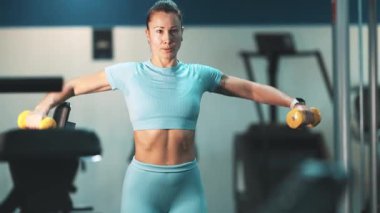 Kas yapan bir kadın, güç antrenmanı sırasında dambıllarla omuz egzersizi yapıyor.