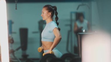 Vücut geliştiren kadın spor salonunda omuz dayanıklılığı için sarı halterlerle yükseltiyor.