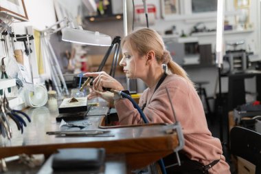 Kadın zanaatkar konsantre oluyor, tezgahındaki metal bir parça üzerinde hassas lehimleme yapıyor.