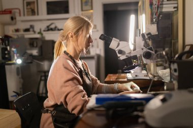 Zanaatkar kadın atölyesinde bir stereomikroskop altında el yapımı bir parçayı dikkatlice inceliyor.