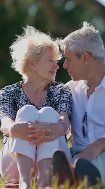 Yaşlı çift parkta battaniyede oturuyor, gülümsüyor ve birbirlerine eşlik ediyorlar.