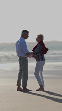 Yaşlı çift gün batımında kumlu sahilde dans ediyor.