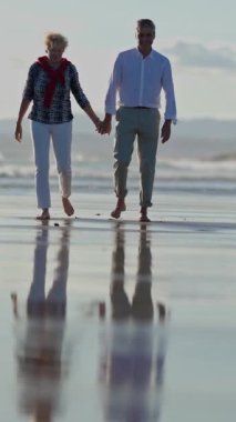 Yaşlı bir çift kumsalda yürümekten zevk alıyor, el ele tutuşuyorlar ve deniz kabuklarını topluyorlar.