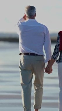 Yaşlı çift kumsalda rahatça yürüyüp el ele tutuşuyor ve emekliliklerinin tadını çıkarıyorlar.