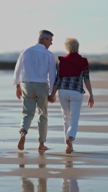Yaşlı bir çift sahilde çıplak ayakla romantik bir yürüyüş yapıyor.