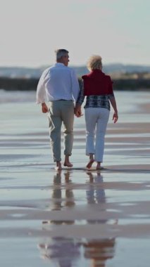 Gün batımında sahilde yürüyen yaşlı çift el ele tutuşuyorlar.