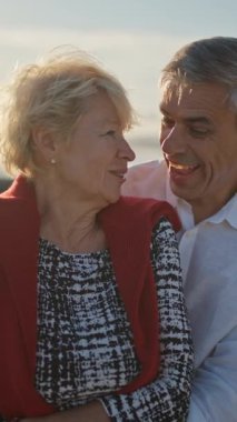 Deniz kenarında romantik bir anın tadını çıkaran yaşlı bir çift güzel bir gün batımında birbirlerine sarılıp gülümsüyorlar.