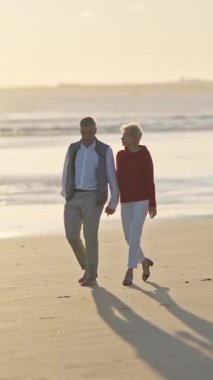 Yaşlı çift gün batımında sahilde romantik bir yürüyüş yapıyor.