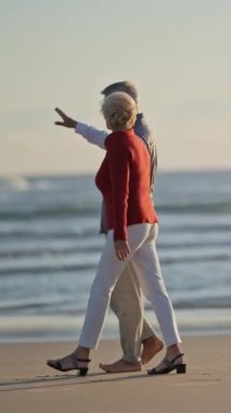 Kıdemli çift gün batımında kumsalda sakin bir yürüyüşün tadını çıkarıyor.