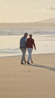 Emekli çift, altın bir günbatımında kumsalda romantik bir yürüyüş yapıyor.