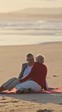 Emekli çift gün batımında battaniyede dinleniyor, okyanus manzarasının tadını çıkarıyor.