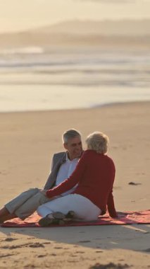 Yaşlı çift kırmızı battaniyede dinleniyor, gün batımında okyanusun kenarında samimi anlar paylaşıyorlar.