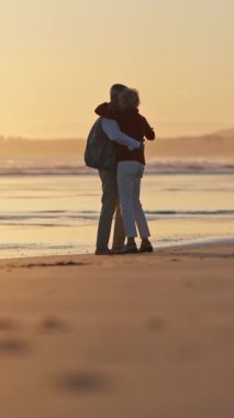 Romantik yaşlı çift gün batımında kumsalda huzurlu bir anın tadını çıkarıyor, kucaklaşıyor ve ufku işaret ediyor.