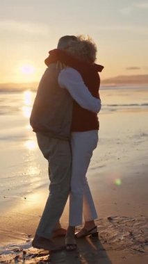 Sevecen yaşlı çift güzel bir gün batımında kumsalda şefkatle sarılıyor.