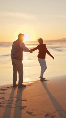 Kıdemli çift el ele tutuşuyor, yürüyor ve gün batımında sakin bir kumsalda kucaklaşıyorlar.