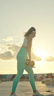 Genç bir kadın, şehir çatılarında günbatımı egzersizleri sırasında dambıl sıralaması yapıyor.