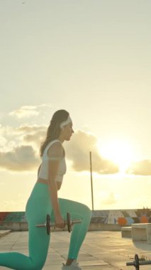 Fitness modeli damda günbatımında dambıllarla hareket ediyor.