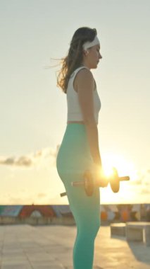Fitness modeli gün batımında damda dambıllarla egzersiz yapıyor.