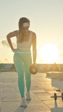 Spor salonundaki kadın gün batımında dışarıda halterlerle egzersiz yapıyor.