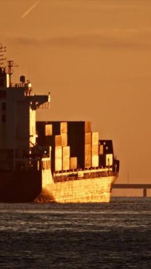 Gün batımında sakin sularda yol alan konteyner gemisi, arka planda görünen bir köprü ile...